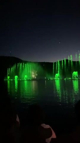 The video displays a nighttime scene of a large-scale water fountain show illuminated by colorful lights. The fountains are situated on a body of water, likely a lake or a large pond, with a dark, hilly landscape in the background. The sky is dark, indicating it is nighttime. In the foreground, the silhouettes of people can be seen watching the spectacle. The fountains spray water high into the air, and these jets of water are illuminated with vibrant green and blue lights, creating a dramatic visual effect that is reflected on the surface of the water. The light colors change throughout the short clip, transitioning from green to various shades of blue.