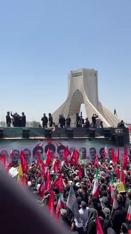 A large, densely packed crowd of people are gathered outdoors, holding numerous red, green, and white flags, many of which bear Islamic imagery and portraits of political figures. In the background, a large, distinctive white monument with an arched opening rises against a clear blue sky. A raised stage is positioned in front of the monument, with a group of people dressed in dark clothing standing on it. Banners featuring portraits of men are displayed behind the crowd, above the stage. The overall impression is of a public gathering or rally.