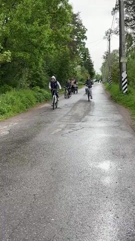 The video shows a group of children and adults on a road lined with trees. Two adults and a child are riding bicycles towards the camera. Behind them, a group of four adults and a small child are walking. The road appears to be wet, suggesting it may have recently rained.  A utility pole is visible on the right side of the road.