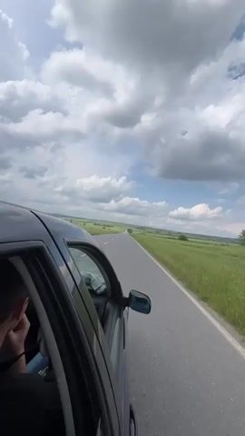 A dark-colored car drives along a straight asphalt road, captured from the back left passenger window. The scene is a rural landscape in Sungurlare, Bulgaria, under a partly cloudy sky with patches of blue. Expansive green fields stretch alongside the road, appearing vibrant and full of tall grass or crops. The car's side mirror is visible in the foreground, and a portion of a person's head and shoulder can be seen inside the vehicle on the left. The video shows the continuous motion of driving through this open countryside on a bright day.