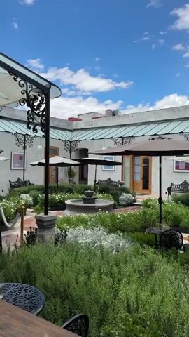 The video presents a view of an outdoor courtyard. The courtyard features a central garden area with a stone fountain and various plants. Around the garden are tables and chairs arranged for outdoor seating. Some seating areas are covered by parasols or a decorative wrought iron structure with a light-colored fabric awning. A tall patio heater stands near the edge of the covered area. The building surrounding the courtyard is white with wooden doors. The sky above is blue with scattered white clouds. No people are visible in the video. The camera pans slightly to show different angles of the courtyard.