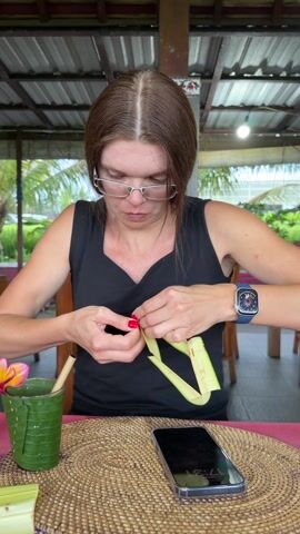 A woman is seated at a table, attentively manipulating strips of green palm leaves, weaving them together. She has medium-length brown hair, wears glasses, and is dressed in a black tank top. Her fingernails are painted red, and she has an Apple watch on her left wrist. She's indoors, under a corrugated iron roof, likely in a restaurant or outdoor dining area, as suggested by the wooden furniture and background scenery with palm trees. A woven placemat anchors the scene on the table, upon which rests a smartphone, a cup made from a palm leaf containing a drink with a flower garnish, and additional strips of palm leaf. The woman is focused on her task of palm leaf weaving, handling the material with precision and care.