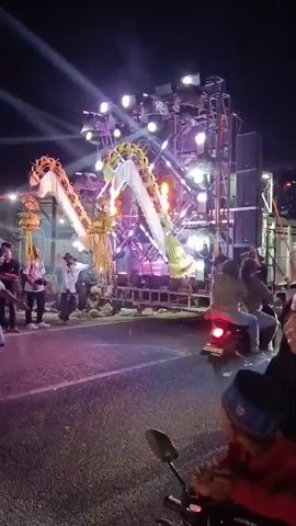 The video captures a nighttime outdoor event, likely a street festival or parade, with a prominent sound system and elaborate light display at its center. The sound system, a large, multi-tiered structure, is equipped with numerous speakers and flashing lights, projecting beams of purple and white light into the darkness. Two large, decorative structures resembling animal heads, possibly giraffes or camels, with shimmering gold accents, are positioned on either side of the sound system. People are scattered around the area, some standing and observing, while others are on motorcycles. In the foreground, two individuals on a motorcycle are visible, with a younger person looking towards the camera. Another person in a black hooded jacket is walking past the motorcycles. The overall atmosphere is festive and energetic, with the lighting creating a dynamic visual experience.