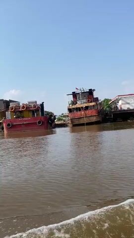 The video shows a body of murky brown water with foamy waves in the foreground. In the background, several large barges and boats are docked along a muddy shoreline. The boats appear to be in various states of repair or disuse, with some showing signs of rust and wear. The barges are large and flat-bottomed, suggesting they are used for transporting goods. The shoreline is lined with trees and some buildings, indicating a populated area. The sky is clear and blue, suggesting a sunny day. The video appears to be filmed from another boat moving through the water, as evidenced by the waves in the foreground.