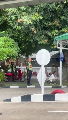 The video captures an outdoor scene in Nong Phueng, Thailand, likely a road safety or driver training area. The time of day appears to be overcast, possibly late morning or afternoon, with diffused lighting.

The primary subjects are several people and motorcycles, suggesting a training or testing environment. In the foreground, a black motorcycle helmet with red accents rests on a railing, implying someone is either preparing to ride or has just finished.

Further into the scene, a man in a bright green vest and black protective gear stands near a red motorcycle, possibly an instructor or examiner. Another person, wearing a similar green vest, is seated on a red scooter to his left, also appearing to be involved in the activity.

To the right, a woman is crouched near a tree, observing. Behind her, a person on a red scooter rides away from the camera. Several other motorcycles are visible in the background, parked or being used. A white sign with a circular element is prominent in the middle ground.

The setting features a covered structure with a green roof, indicative of a waiting or briefing area. The ground is paved, with black and white striped curbs and some red tires used as markers, common in training courses. Lush green trees and foliage surround the area, typical of a tropical climate. The presence of multiple individuals and motorcycles in what seems to be an organized setup points towards a motorcycle riding lesson or test. No specific emotions are discernible, but the scene conveys a sense of practical training or assessment. There is some text on a blue sign near the covered structure, but it is not legible.