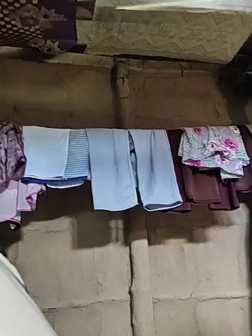 The video shows a clothesline hanging horizontally across a rough, earthen wall. On the clothesline are several items of clothing, including a patterned purple shirt on the far left, a light blue towel, a striped blue towel, and two light blue pants. To the right of the pants are a dark brown garment and a floral-patterned shirt. The environment appears to be indoors, with evidence of a ceiling or overhead structure visible at the top of the frame. The wall is textured and uneven, suggesting it may be made of mud or a similar material. A blurred white object is visible in the lower left corner of the frame. The lighting is dim, creating shadows on the wall behind the clothesline. The video is static with no visible movement.