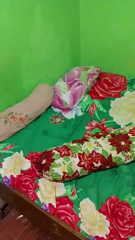 A camera slowly pans across a bed covered in a green floral comforter. The comforter is decorated with large red, yellow, and purple roses, along with smaller red and white poinsettia-like flowers. Three pillows are visible on the bed. One is a long bolster pillow with a poinsettia pattern. Another is a square pillow with a pink and green floral design. A third, lighter colored pillow is partially visible behind the bolster. In the lower right corner of the frame, a folded garment, possibly a sarong or traditional clothing, is placed on the bed. The wall behind the bed is painted a bright green. The lighting is dim, suggesting it may be indoors or at night. The camera movement is smooth and steady, capturing the textures and patterns of the bedding.