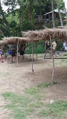 The video shows a group of people at a river or swimming hole.  Some people are standing under several rustic, thatched-roof structures that appear to be shelters. Others are standing near a rocky area, and a few are on or near a large rock in the water. There are trees and other foliage around the river, with a small building visible higher up on the landscape.