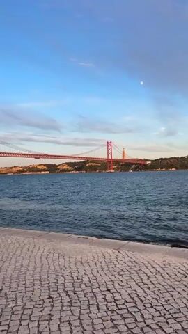 The media file captures a serene waterside view of the 25 de Abril Bridge in Lisbon, Portugal, during what appears to be sunset or early evening. The sky is a gradient of blue with wisps of pink and white clouds, and a pale moon is visible. The iconic red suspension bridge stretches across a wide, dark blue body of water, likely the Tagus River.

In the foreground, a textured cobblestone walkway lines the edge of the water. The water itself is calm with gentle ripples. On the far bank, beyond the bridge, a lush green hillside rises, topped with trees and buildings, including a prominent statue that is characteristic of Lisbon's Christ the King monument.

The scene is tranquil, bathed in the soft light of the setting sun. There are no people visible in the frame, emphasizing the peaceful, natural beauty of the location. The overall mood is one of calm and quiet contemplation, inviting the viewer to appreciate the scenic landscape. The colors are rich and warm, reflecting the time of day. There is no visible text in the media file.