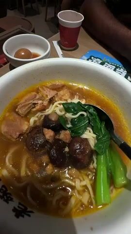 The video shows a close-up view of a bowl of noodles, likely beef noodle soup, with chunks of meat, mushrooms, and green vegetables. A spoon is seen stirring the contents of the bowl. In the background, there is a small white bowl with two brown eggs and some sauce, a red and white disposable cup, and a wooden table with some papers. The lighting suggests an indoor setting, possibly a restaurant. The perspective is from above, looking down into the bowl.