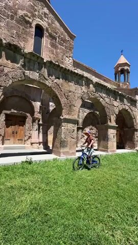A young person on a blue bicycle rides on a grassy lawn in front of a stone building with arched doorways. The sky is clear and blue.
