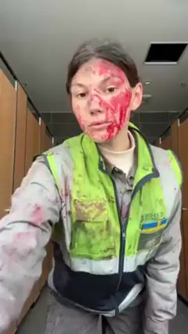 A person stands in a public restroom, covered in red stains resembling blood. The individual is wearing a high-visibility work jacket with the Swedish flag patch, gray pants, and a gray shirt. The blood-like stains cover the person's face, arms, and pants. The bathroom has multiple closed stall doors on either side of the person, a tiled ceiling, and bright overhead lighting. The person stands still and looks at the camera with their hands in their pockets.