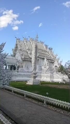 The video shows a wide shot of a large, ornate white temple complex under a blue sky with scattered clouds. In the foreground, a pathway with a decorative white railing leads towards an intricately carved white archway. Beyond the archway, the main temple building is visible, characterized by its multi-tiered roof and elaborate white ornamentation. A single person, dressed in dark clothing, stands near the entrance of the archway, appearing to walk into the temple complex. The surrounding area consists of a green lawn.
