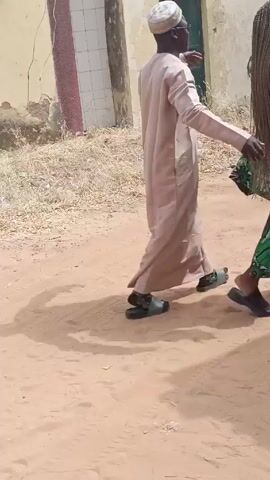 A man in a long tan robe and white cap walks across a sandy, dirt area. He is wearing dark sandals. To his right is a woman in a green and patterned dress. They are near a building that has a green door. There is straw or dried grass on the ground. The man gestures with his right arm.