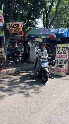 The video shows a young male sitting on a scooter in front of an outdoor food stall. The stall has signs for "Bakso Mie Ayam" and "Es Teh Jumbo" and "Es Jeruk Jumbo." The young male is wearing a light-colored long-sleeved shirt and brown pants. He is looking towards the camera. Behind him, there are other stalls, people working, and trees. The lighting suggests it is daytime, likely sunny. The ground is paved.