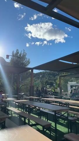 The camera pans across an outdoor beer garden, focusing on people and the surroundings. The scene is filled with rows of wooden picnic tables arranged under a large, shaded canopy. The blue sky with scattered clouds is visible above the canopy. People are seated at some tables, chatting and enjoying their time. In the background, there are booths with signage. The scene is situated in a mountainous setting, with trees and rock formations visible in the distance. The camera captures various individuals, including a woman wearing a colorful headscarf, a man in sunglasses, and groups of friends interacting.