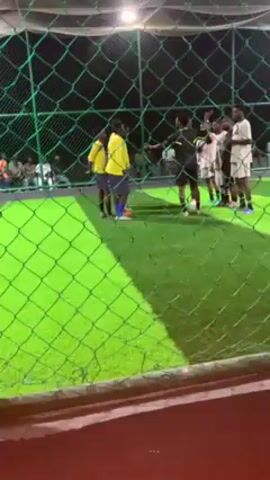 The video shows a soccer game at night, viewed through a green mesh fence. Two teams, one in yellow and one in white and black, are gathered around the ball, presumably discussing strategy or addressing a rule. A referee in black is also present, seemingly mediating the discussion. The field is artificial turf, brightly lit by overhead lights. Several people are seen watching from the sidelines. The camera view is partially obstructed by various individuals wearing different clothing near the fence.