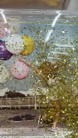 The video shows a clear glass container with a liquid, possibly water, inside. A print of a woman holding a bunch of balloons floats within the liquid. Gold glitter is suspended in the liquid, appearing to be falling. The print depicts the woman in a white dress with a black top, running with her hair flowing, as if being lifted by the balloons. In the background of the print, there is a cityscape with buildings and a bridge or fence. The glitter creates a festive, celebratory atmosphere around the image.