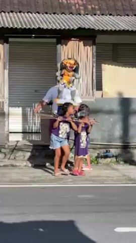A man and two young girls are carrying a statue of a demon along a road in a residential area. The man, dressed in a white shirt and purple wrap, carries the front end of two parallel wooden poles. The two girls, wearing matching purple shirts and denim shorts, carry the back end. The statue, on a blue and yellow platform, depicts a demonic figure with a sword. The houses in the background are simple, with tiled roofs and some greenery. Another woman in a white patterned dress is seen briefly behind the group.