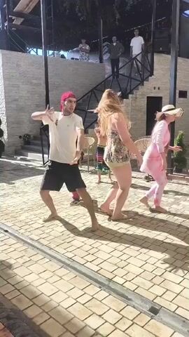 Four individuals are dancing outdoors on a paved patio. On the left, a person in a red and black Deadpool costume is dancing. Next to them, a woman in a floral bikini is dancing. To her right, a man in a white t-shirt and black shorts is dancing barefoot. On the far right, a woman in a pink tie-dye outfit and a straw hat is dancing. In the background, several other people are visible on a raised platform with stairs. The scene appears to be during the day, with sunlight casting shadows.