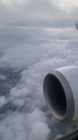The view from an airplane window shows a white jet engine against a background of dense clouds. The sky is overcast with a hint of sunlight peeking through the clouds in the upper portion of the frame. The clouds appear layered and extend to the horizon. The turbine is static, suggesting a stable flight.