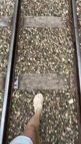 The media file shows a first-person perspective of walking along train tracks. The subjects are a person's legs and feet wearing a sandal. The scene is the train tracks themselves, with gravel ballast and wooden ties. The setting appears to be a natural environment with trees lining the path, and a wooden fence alongside the tracks. In the distance, there are benches and lampposts, suggesting a developed pathway or station area.

The activity is walking. The time of day appears to be daytime, given the lighting. The weather seems fair, as there is ample natural light and no visible precipitation. The location cues, such as the style of the lampposts and the surrounding foliage, might hint at a specific region, but without more information, it's difficult to pinpoint precisely. However, the provided context mentions Guamka, Russia. The overall mood conveyed is one of exploration or travel. There are some blue flags with text on them visible in the distance, but the text is not legible.