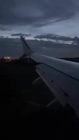 The video shows a view from an airplane window during landing at night. The wing of a Ryanair airplane is visible with the airline's name on its fin. The landing strip with green and red lights is seen below as the plane approaches. The sky above is dark with clouds. Beyond the runway, distant city lights twinkle.