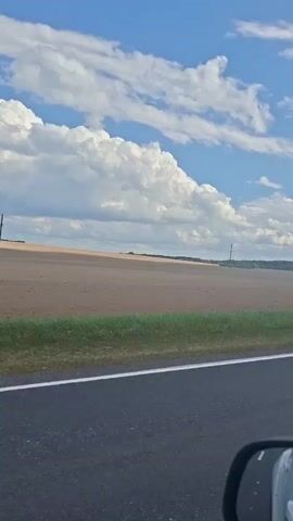 THE VIDEO SHOWS A VIEW FROM A MOVING VEHICLE. THE FOREGROUND FEATURES AN ASPHALT ROAD WITH A WHITE LINE ON THE LEFT. TO THE RIGHT OF THE ROAD, THERE IS A GREEN GRASSY STRIP BORDERING A LARGE, FLAT, BROWN FIELD. THE FIELD APPEARS TO BE CULTIVATED OR HARVESTED. IN THE BACKGROUND, THE FIELD CONTINUES TO A HORIZON WHERE TREES ARE VISIBLE. TWO TALL, THIN POLES, LIKELY UTILITY POLES, ARE DISTANTLY SEEN STANDING IN THE FIELD. THE SKY IS BLUE WITH LARGE, PUFFY WHITE CLOUDS. THE REFLECTION OF THE VEHICLE'S INTERIOR, INCLUDING A REARVIEW MIRROR, IS VISIBLE IN THE BOTTOM RIGHT CORNER OF THE FRAME. THE VIDEO APPEARS TO BE CAPTURED DURING DAYLIGHT HOURS.