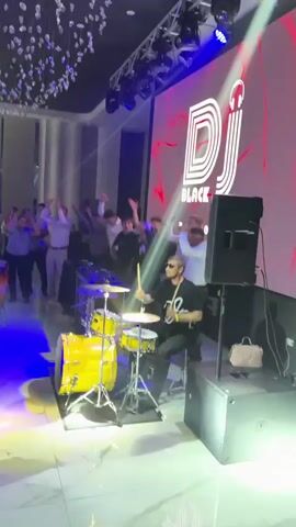 A large group of people are dancing and celebrating in a dimly lit room, possibly a ballroom or event hall, decorated with white chandeliers and spotlights that cast blue and white light. In the foreground, a drum set with yellow drums is visible. A person is seated behind the drum set, seemingly playing. To the left, a conga drum is also present. A large screen at the back of the room displays the words "DJ BLACK" in white lettering. Several people are standing and raising their hands in the air, some are in motion, dancing. The atmosphere appears festive.
