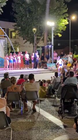 An outdoor nighttime event is taking place in front of a stage. Several people are performing on the stage, some appear to be dancers in brightly colored outfits, and a few are standing behind microphones. A crowd of people is seated and standing in front of the stage, watching the performance. There are trees, streetlights, and buildings in the background. Some people in the crowd are holding what appear to be cell phones, possibly recording the event.