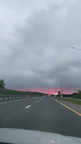 The video shows a view from inside a vehicle traveling on a highway. The sky is overcast with dark gray clouds, but a band of pink or red light is visible near the horizon, suggesting a sunset or sunrise. The highway has multiple lanes with white and yellow markings. There are guardrails on the sides of the road, and some vegetation is visible beyond them. Several cars are driving in the distance. The road surface appears wet in some areas, with visible reflections. The camera is positioned as if looking forward from the dashboard or windshield.