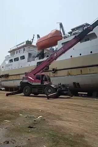 A large pink and black crane is positioned next to a docked white and yellow ship. The crane's arm extends upwards, lifting an orange lifeboat. The ship is large, with multiple decks and railings visible. The environment appears to be a dry dock or shipyard, with a dirt ground and other machinery or vehicles present in the background, including a red truck. The sky is overcast. The crane has the logo "ISSEY" on its arm. The crane is labelled "CRN 1". The lifeboat is suspended by the crane's hook. The ship's hull appears weathered and rusty in some areas. The action captured is the removal or placement of the lifeboat using the crane.