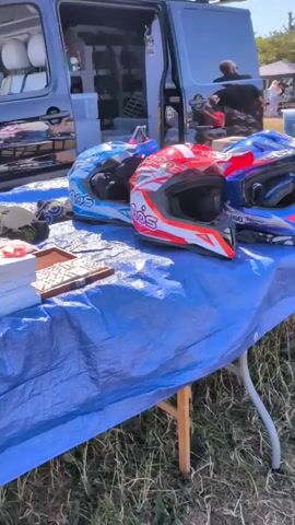 The video shows a close-up of several motorcycle helmets displayed on a blue tarp spread over a table. The helmets are of various designs and colors, including a red and white motocross helmet, a blue and red motocross helmet, and two black helmets with white stripes. In the background, a black van with its doors open reveals storage containers and some items inside. There are other people and objects visible in the distance, suggesting an outdoor event like a market or a race. The lighting indicates it is daytime.