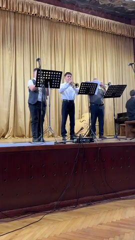 Three male musicians perform on a wooden stage with golden pleated curtains as a backdrop. Three men are standing in the foreground, each playing a trumpet and reading from sheet music on individual stands. Two microphones on stands are positioned in front of the trumpeters, suggesting amplification of their instruments.

The man on the left wears a white shirt and a dark vest, with black pants. The central musician, also in a white shirt and black pants, plays his trumpet while facing forward. The man on the right wears a grey polo shirt and dark pants, also playing a trumpet.

Behind them, an upright bass is visible. Further to the right in the background, a fourth musician is seated on a wooden stool, partially obscured by the trumpeters, and appears to be playing a piano.

The stage itself has a dark brown front panel and a lighter wooden floor. Black cables are visible on the stage floor, connecting to various equipment, including a small black device with the word "INVOTONE" printed on it. The floor in front of the stage is a wooden parquet pattern. The overall scene depicts a live musical performance.