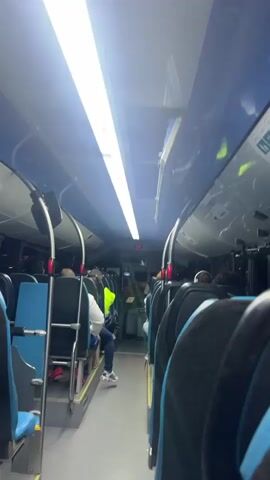 The video captures the interior of a moving bus or coach, featuring rows of blue and black seats. Overhead, bright white fluorescent lights illuminate the cabin. Several passengers are visible, mostly seated; one individual is wearing a bright yellow vest. The main focus of the video is an overhead compartment that has opened, revealing light blue insulation material protruding from it. A digital display shows "2 08". The ambient lighting suggests it is dark outside, possibly nighttime. The event takes place in Telde, Spain.