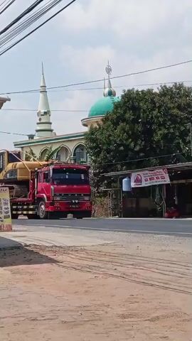 A red flatbed truck carries a yellow excavator on its trailer. The truck is driving on a road next to a mosque with a green dome. Trees and power lines are visible in the background. The excavator is a Caterpillar brand. The scene appears to be in a rural or suburban area during daylight.