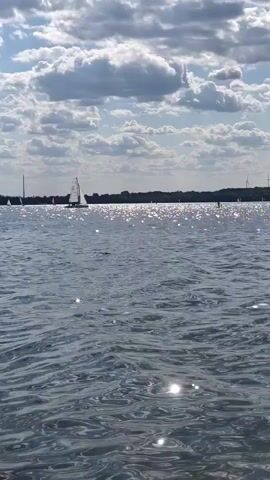 The video shows a wide shot of a lake on a sunny day with scattered clouds. The water is rippling, and sunlight glints off its surface. In the distance, a sailboat with white sails is moving across the water. The shoreline is visible in the background, with trees lining the horizon. There are no people visible in the foreground, only the expansive view of the lake and sky.