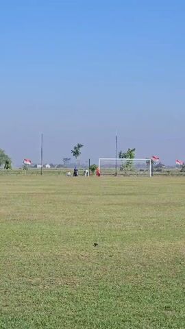 A wide shot of a grassy soccer field under a clear blue sky. Indonesian flags are mounted on poles along the perimeter of the field. A soccer goal stands on the far side of the field, with a few figures near it, one wearing red. The grass appears well-maintained. The atmosphere is serene and rural.