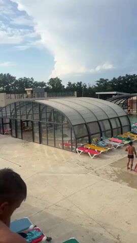 The video shows an outdoor water park on a cloudy day. In the foreground, a person wearing dark shorts walks away from the camera. To the left, two enclosed swimming pool structures with arched, transparent roofs are visible. Deck chairs with colorful cushions are arranged in rows outside these structures. In the background, two water slides, one yellow and orange, the other green and blue, are prominent. Several people are seen on the slides and in the water. A lifeguard in a yellow shirt and red shorts sits on a raised chair near the slides. The environment is paved with concrete, and trees line the background.