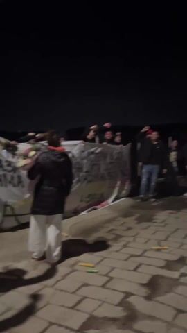 At night, a group of people stand behind a large banner on a brick-paved surface. Most are young men. Many raise their fists in the air and appear to be chanting. A drummer stands to the right, playing a large, white drum. A person in a dark jacket and white pants walks into the frame. The banner contains writing in a language other than English. Some discarded items, possibly noisemakers, are scattered on the ground. The environment appears to be outdoors.