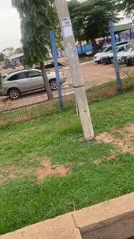 The video pans across a scene in Kinkinau, Nigeria, likely a perimeter or checkpoint area. In the foreground, a concrete structure with exposed black and white cables runs alongside a grassy area. Beyond a chain-link fence, several cars are parked. A tall, weathered pole stands prominently, with a sign labeled "ZONE 10 16" attached to it. In the background, a small, blue and white building with a green roof is visible, and a pathway with yellow and black striped bollards and a chain suggests a controlled entry or exit. The overall atmosphere appears neutral, possibly during daylight hours with overcast skies. No people are visible, and the focus is on the infrastructure and setting.