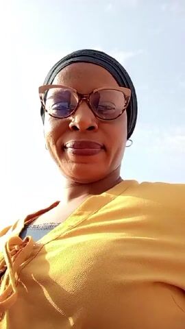 A woman wearing a yellow shirt, a black head covering, and glasses looks directly at the camera. She is outdoors with a white sky overhead. The camera angle is low, looking up at the woman. In the background there is a very loud noise, potentially from a jet engine. She makes a slight pursing of the lips, creating a small kiss-like expression.