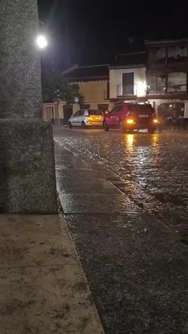 The video, filmed at night, shows a wet cobblestone street with two cars. A light-colored sedan is parked on the left, and a red SUV is parked on the right, with its brake lights illuminated. The street is reflecting the lights from the vehicles and streetlights. Buildings with balconies and lights are visible in the background. It appears to be raining, as indicated by the wet surfaces and possible raindrops visible in the air.
