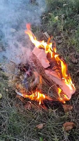 A bundle of dried plants and paper is burning on the ground, surrounded by grass and fallen leaves. Flames engulf one side of the bundle, while smoke billows from the other. The fire is intense, with bright orange and yellow flames. The scene appears to be outdoors, in a natural setting. The camera is positioned above and to the side of the burning bundle, providing a clear view of the action.