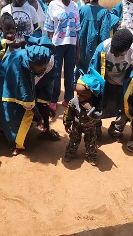 The video captures an outdoor gathering, likely a community event or celebration, with several individuals, predominantly children and young adults, dressed in teal robes with yellow trim. The ground appears to be dirt or sand, suggesting a rustic setting. In the foreground, a small child in a grey and camouflage-patterned outfit, topped with a teal cap, is interacting with an adult also wearing the teal robe. The adult is bent down, gesturing towards the child, who is looking down and touching their mouth. Several other people in similar robes are standing and observing, some with smiles on their faces. The atmosphere appears cheerful and engaged, with multiple people looking towards the central interaction. The lighting suggests it is daytime, with shadows cast on the ground, indicating sunlight. The overall scene conveys a sense of communal gathering and celebration, possibly a graduation or religious ceremony, given the attire.