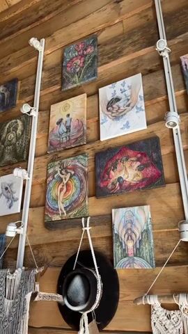 The video shows a wooden wall decorated with several paintings.  A white track lighting system runs vertically on the wall. The paintings depict various subjects, including flowers, figures in natural settings, and abstract designs. A black hat hangs below the paintings. The wall appears to be part of a rustic-style interior.