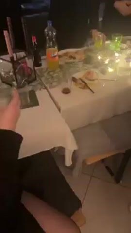 The video shows an indoor gathering of approximately 8-10 people. Some are standing around a table laden with food and drinks, including a Fanta bottle. There are decorative candles and holiday themed decorations on the table. People are dressed in both casual and semi-formal attire. At least one person is recording with their phone. The video shows multiple rooms, including one with a bar. There is a mirror reflecting the gathering. The video appears to be shaky and the focus shifts often.