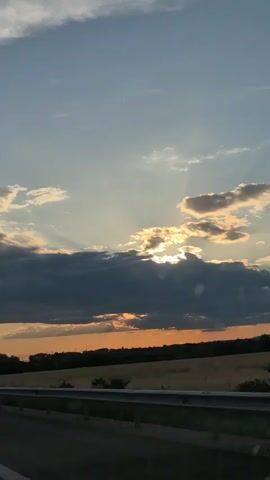 The video shows a view from a moving vehicle looking out at a cloudy sky during sunset. The sun is partially obscured by dark clouds, but rays of light are breaking through, illuminating the sky. Below the sky is a horizon line with trees and fields. A guardrail runs along the side of the road in the foreground. The overall scene is one of natural beauty during twilight.
