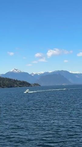 The video shows a serene view of a mountain range covered in snow.  The sea in the foreground is dark blue, contrasting with the clear blue sky dotted with puffy white clouds. A small white boat cuts through the water, leaving a white wake. The scene appears calm and peaceful.