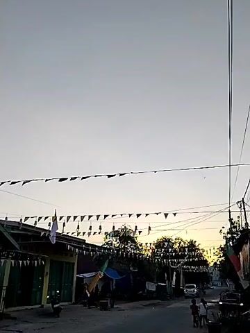 The video captures a street scene with small buildings lining both sides. Decorative flags are strung across the street, creating a festive atmosphere. In the foreground, a dirt road runs through the middle of the frame. Several small shops or houses are visible, with some appearing to be closed. A few people are seen walking along the street. The sky is clear and overcast, suggesting it might be early morning or late afternoon. The overall impression is of a quiet, rural or semi-urban setting decorated for an event.
